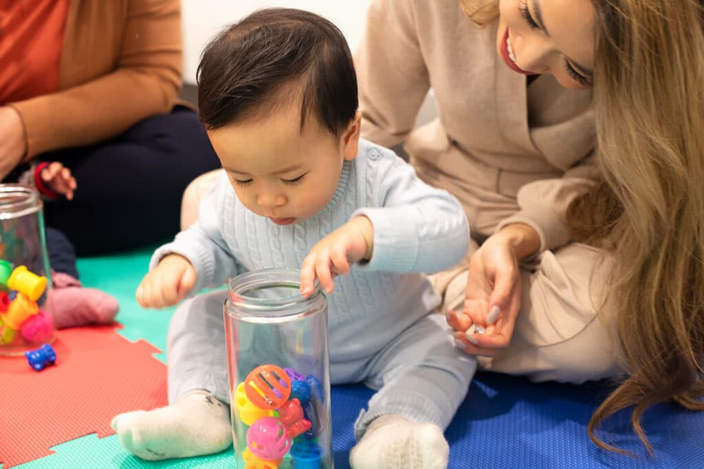 Fine motor skills development, A mother and her baby sitting on a colorful play mat engaging in sensory activities with clear jars and colorful plastic toys.