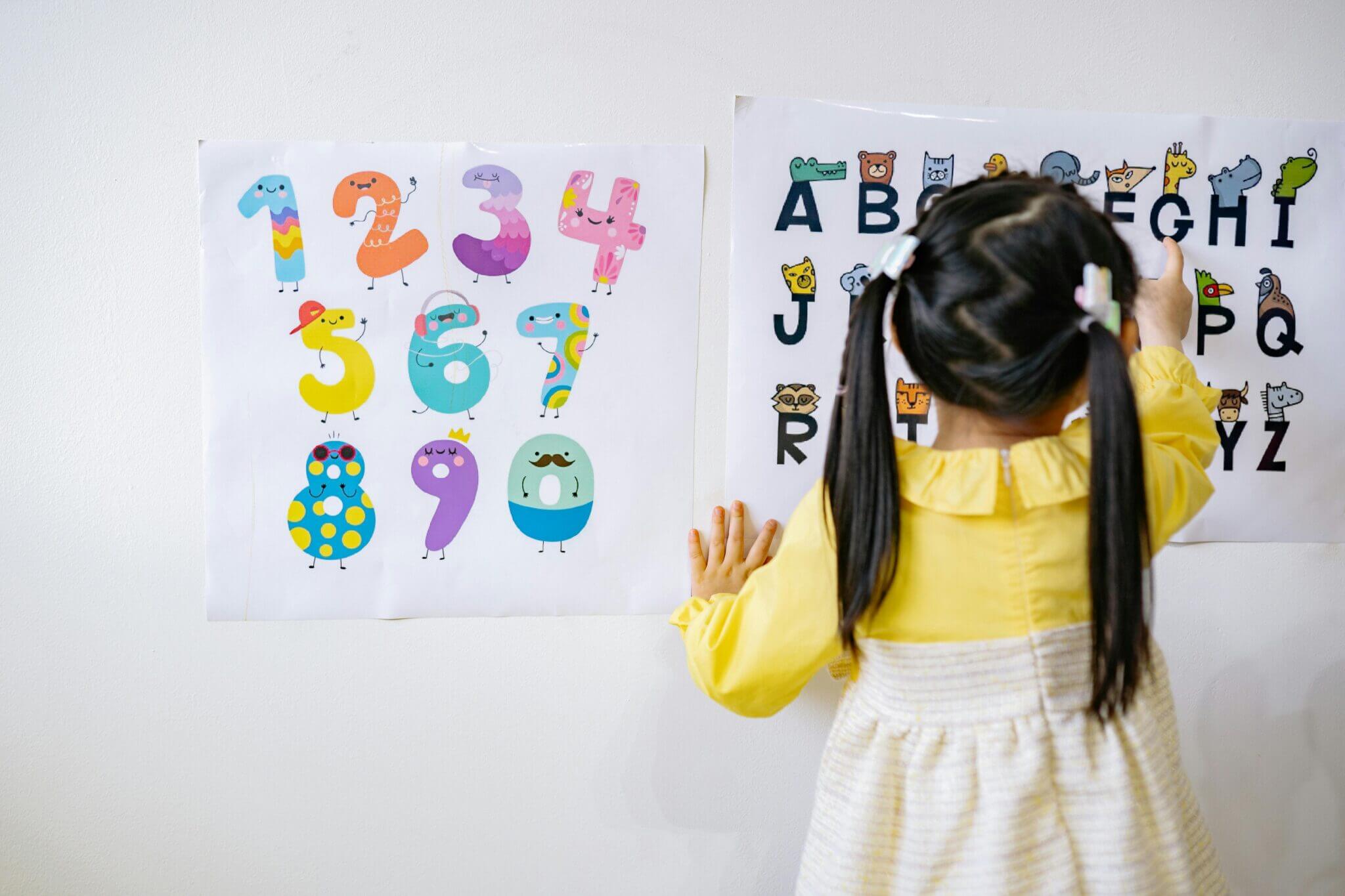 Young girl learning letters with an alphabet poster on a wall. Learning letters and numbers early is part of the Shichida Method.