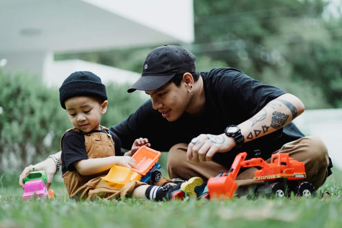 A father plays with his young son outside, they are sitting on the grass playing with toy vehicles.