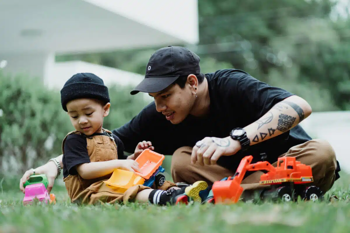 A father plays with his young son outside, they are sitting on the grass playing with toy vehicles.