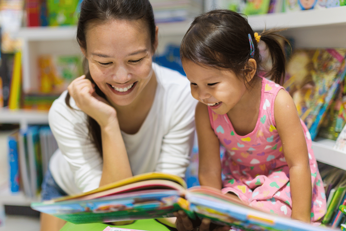 A mum and her young daughter reading together in a library, looking happy