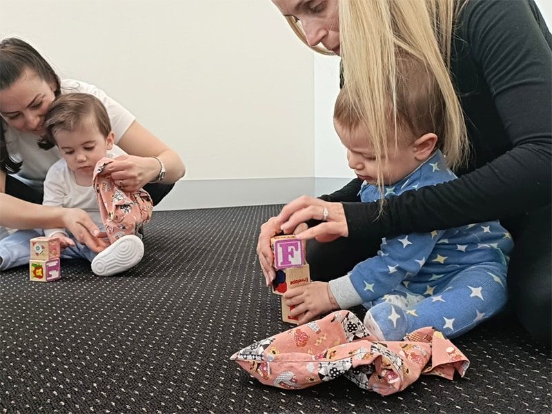 Babies taking part in a block stacking activity with their mums, they are sitting on a carpet and practising together, developing fine motor control, spatial awareness and cause and effect.