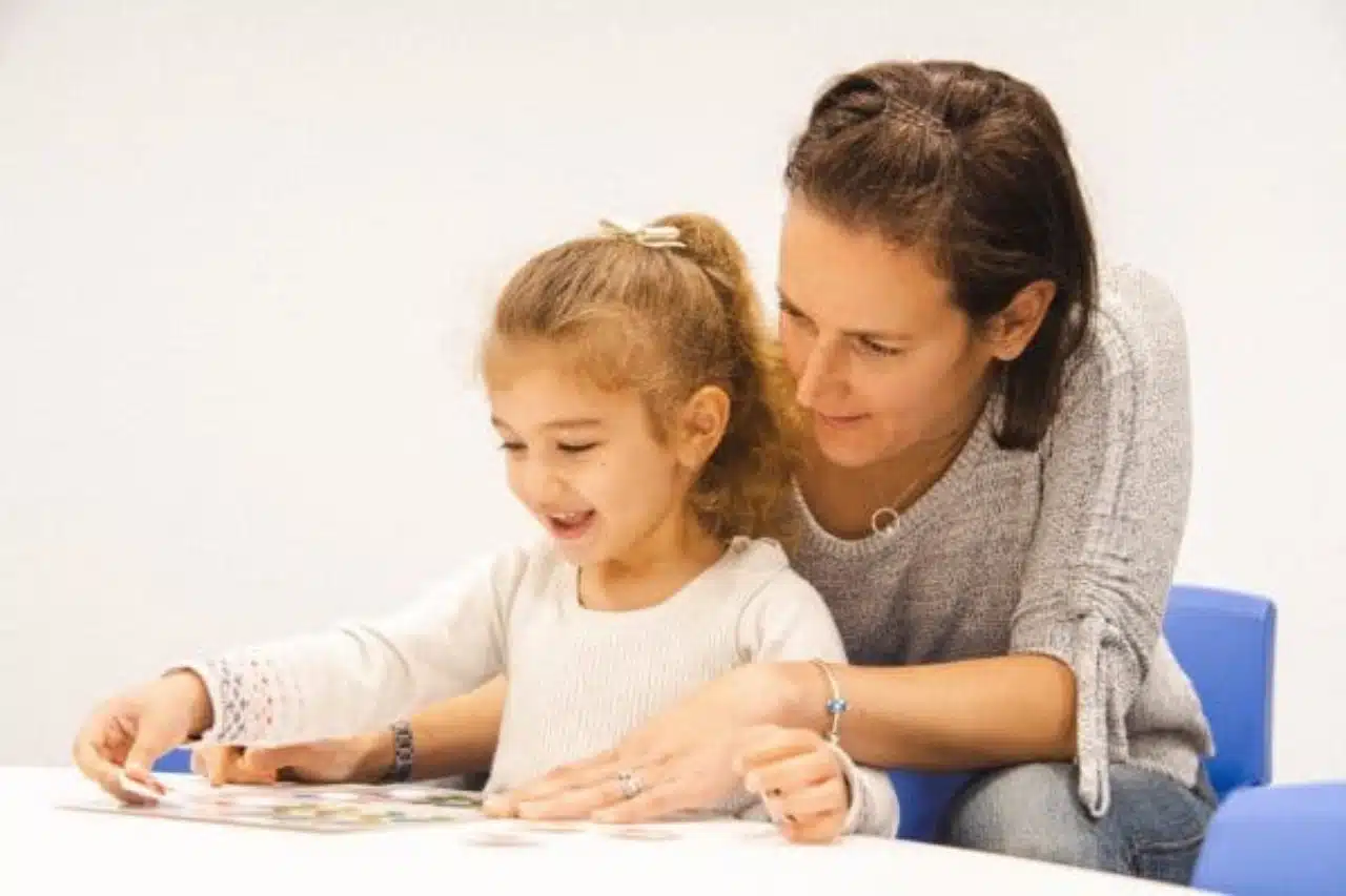 A woman and a child sit at a table, reading a book together, sharing a moment of learning and connection