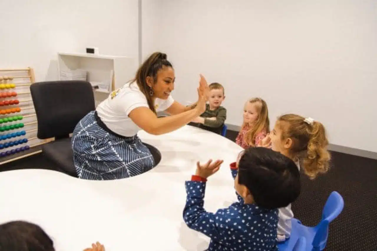 A teacher engages with children in a classroom setting, surrounded by learning resources and student artwork on the walls
