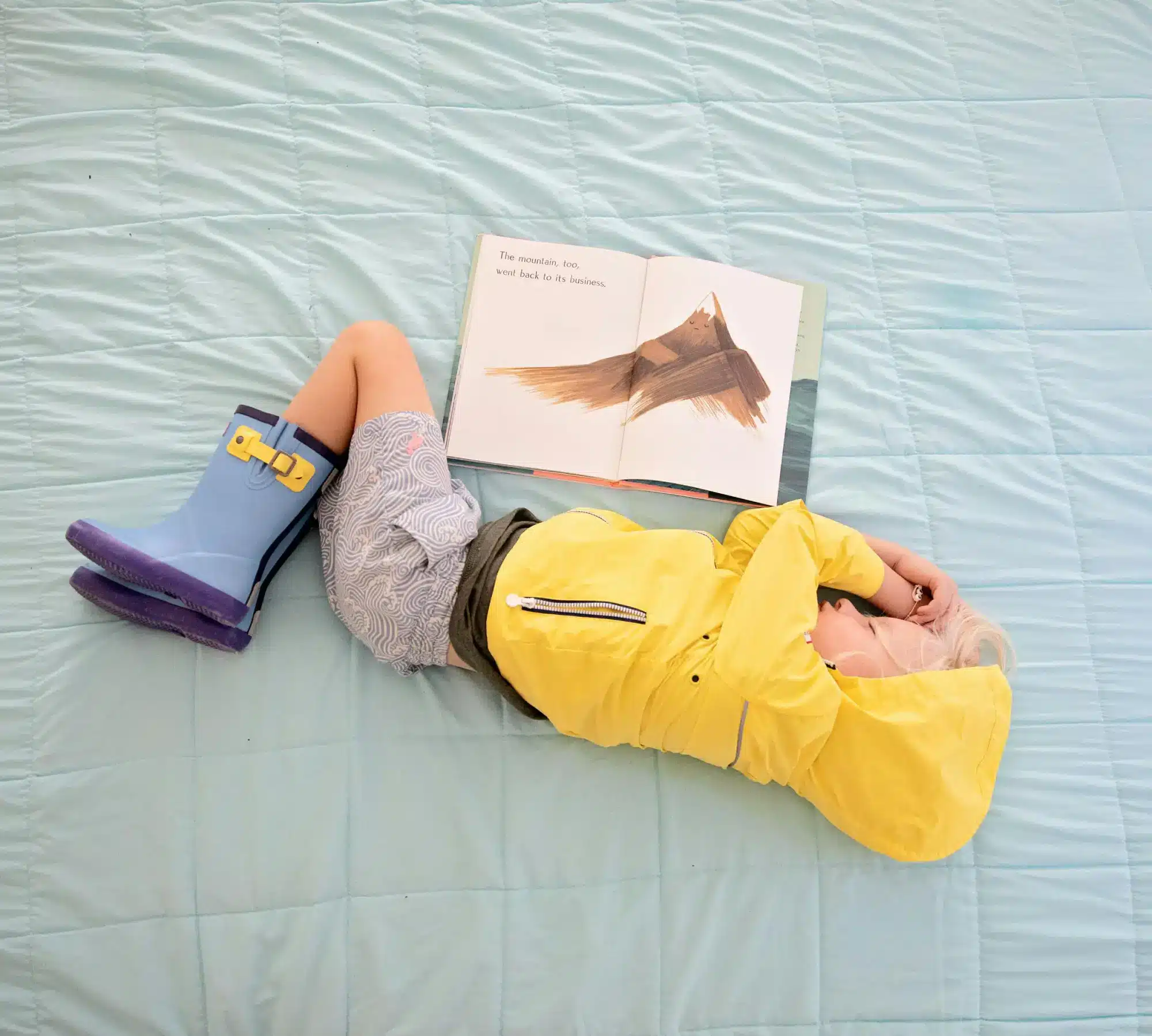 A young child in a yellow raincoat and blue rain boots lies on a blanket with an open book in front of them.
