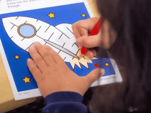 A child's hands holding a red pencil and tracing a rocket ship on a sheet of paper.