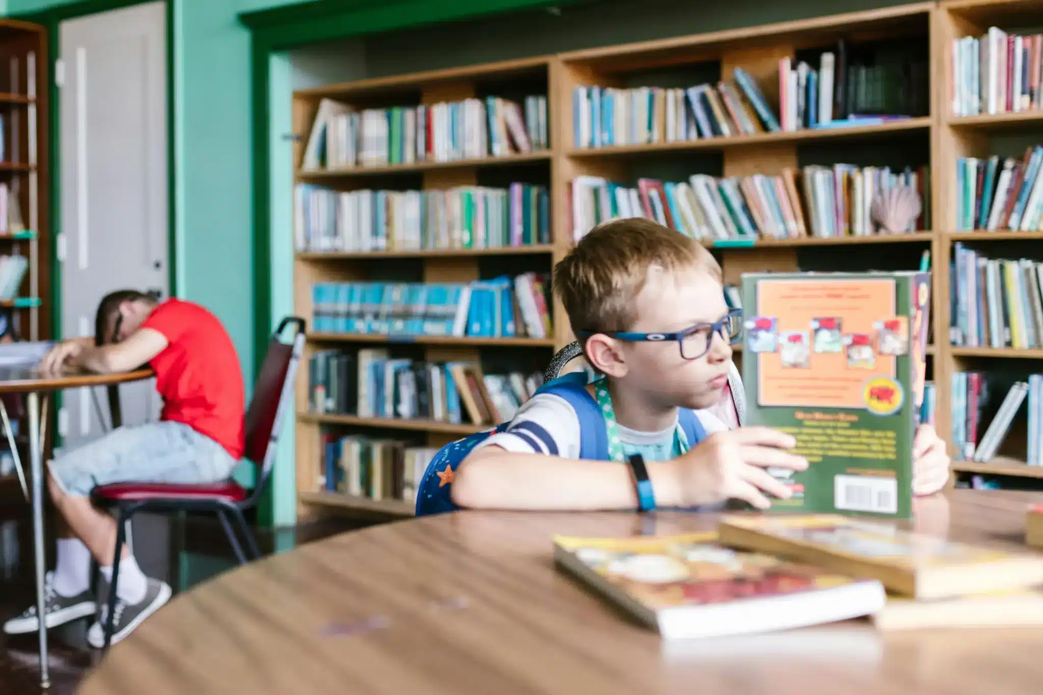 A young boy with glasses sitting at a table in a library, holding a book, while another boy is slumped over a table in the background.