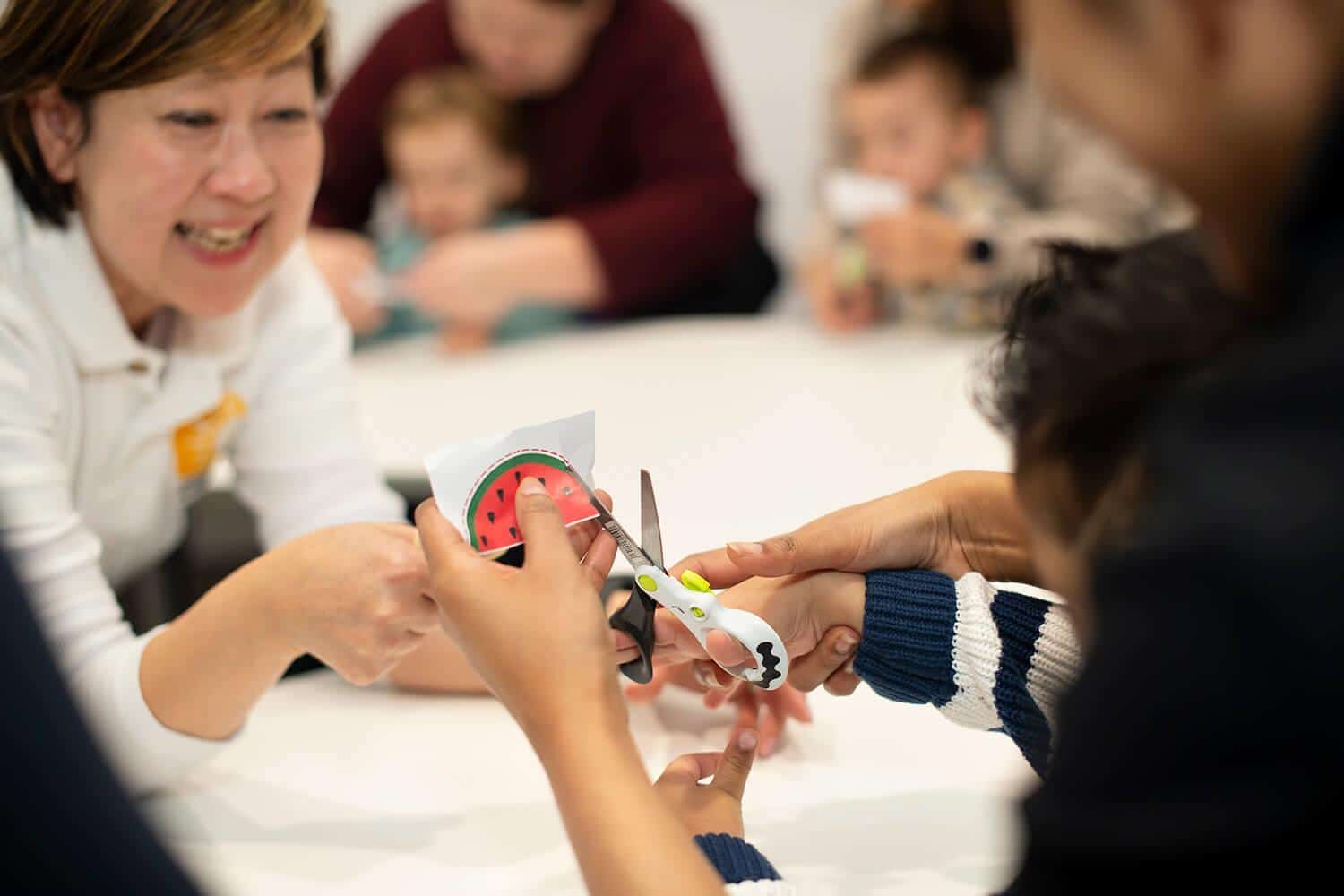 Shichida Australia, a teacher and a parent guide a toddler to cut a curved edge with a safety scissor