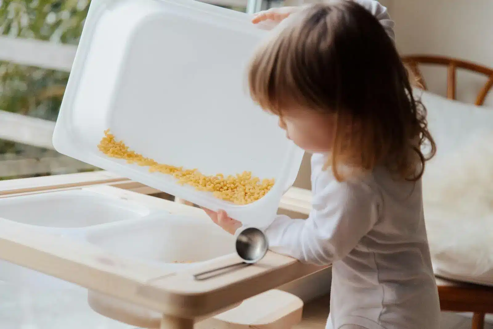 Sensory play for babies. A young child in a white shirt stands at a wooden sensory table, tipping a large white tray to pour dry, yellow pasta into one of the table's white basins.